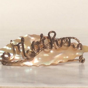 May include: A gold wire name brooch with the word "Mother" spelled out on a pearlescent shell.