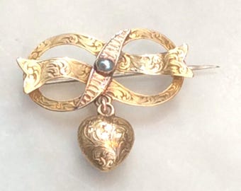 May include: A gold-toned brooch with a heart-shaped pendant. The brooch is in the shape of two intertwined ribbons with a small pearl-like stone in the center. The heart pendant is detailed with an engraved floral pattern.