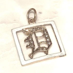 Op de afbeelding: Een gouden bedel met een wit emaille frame en een met parels bezette letter D.