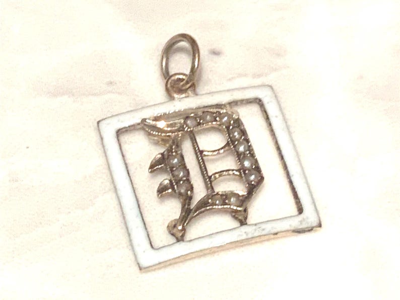 Op de afbeelding: Een gouden bedel met een wit emaille frame en een met parels bezette letter D.