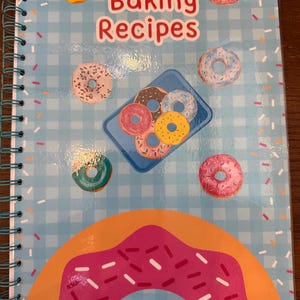 May include: A spiral-bound notebook with a light blue and white checkered cover featuring the text Baking Recipes in red. The cover is decorated with various colorful donut illustrations, including a large pink frosted donut at the bottom.