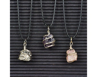 Puede incluir: Tres collares con colgantes de cristal envueltos en alambre. Los colgantes son de amatista, cuarzo transparente y cuarzo rosa. Los collares están sobre un fondo negro.