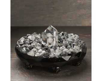 May include: A black crystal altar with a clear quartz point in the center. The altar is surrounded by smaller clear quartz crystals and black stones.