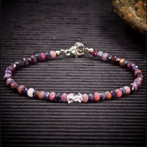 May include: A delicate bracelet made with small, faceted gemstones in shades of blue, pink, and red. The bracelet features a silver clasp and a single clear crystal bead.