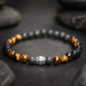 Puede incluir: Un brazalete con cuentas de ojo de tigre marrón y cuentas de lava negra, separadas por una cuenta plateada con un diseño geométrico.