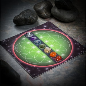 May include: A set of seven chakra stones arranged in a line on a green and black card with a flower of life design. The stones are clear, purple, blue, green, yellow, orange, and red.