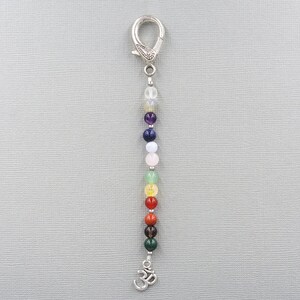 May include: A beaded charm with a silver clasp and a dangling Om symbol. The charm features a string of colorful, round beads in various colors, including green, red, orange, yellow, pink, purple, and blue. The beads are strung together, creating a vibrant and eye-catching accessory.