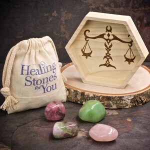 May include: A wooden hexagonal tray with a Libra zodiac symbol engraved on it. The tray is sitting on a wooden slice. There are three tumbled stones in front of the tray: a pink rose quartz, a green aventurine, and a pink and green rhodochrosite. A small white drawstring bag with the text "Healing Stones for You" is in the background.