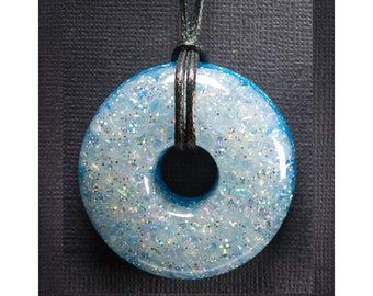Op de afbeelding: Een blauwe en witte glitterhars donut hanger aan een zwarte koordsnoer.