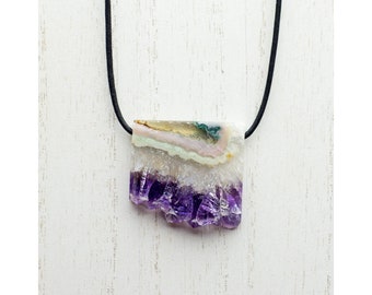 Puede incluir: Un collar con un colgante de cristal de amatista púrpura con un cordón negro. El cristal es en bruto y tiene una forma natural irregular. Tiene una capa blanca y verde encima de la amatista púrpura.