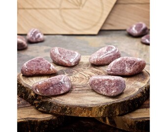 Può includere: Una collezione di otto pietre di lepidolita rosa levigate su una superficie di legno. Le pietre sono lisce e hanno un colore rosa chiaro con striature bianche.