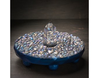 May include: A clear crystal point sits atop a blue circular base, surrounded by iridescent glass stones. The base has three rounded feet.