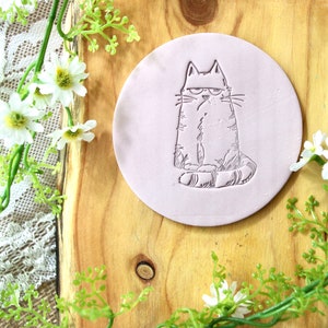 May include: A light pink round coaster with a black and white line drawing of a cat with a grumpy expression. The cat is sitting with its tail wrapped around its legs.
