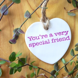 以下が含まれることがあります： 木製の背景に緑色のつるが絡まっている中で、麻ひもで吊るされた白いハート型のセラミック製のオーナメント。オーナメントには紫色の文字で「You're a very special friend」と書かれている。