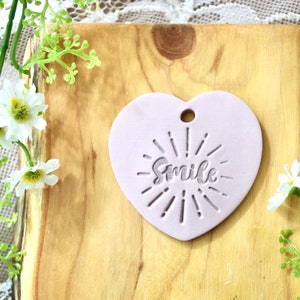 Könnte beinhalten: Ein helllilafarbenes, herzförmiges Keramik-Ornament mit dem Wort "Smile" in einer stilisierten Schrift und einem Sonnenstrahlen-Design.