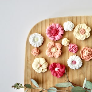 May include: Assortment of 12 small, decorative, sugar paste flowers in various shades of pink, peach, and white. The flowers are arranged on a wooden surface.