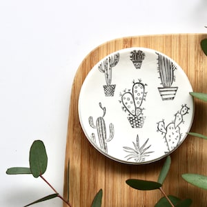 May include: A white ceramic dish with a black and white cactus design. The dish is on a wooden surface.