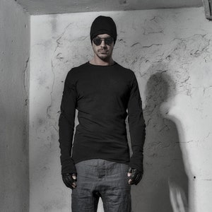 May include: A man wearing a black beanie, black sunglasses, a black long-sleeved shirt, gray shorts, and black shoes. He is standing in front of a white wall.