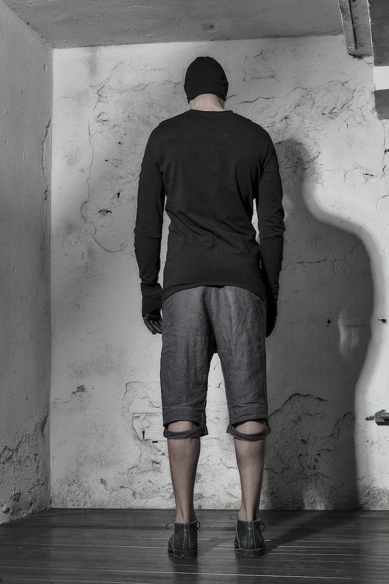 May include: A person wearing a black long-sleeved shirt, black shorts with knee pads, and black shoes. The person is standing in a room with a white wall.