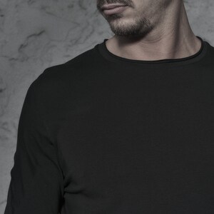 May include: A man wearing a black beanie and black sunglasses with round lenses. He is wearing a black long-sleeved shirt.