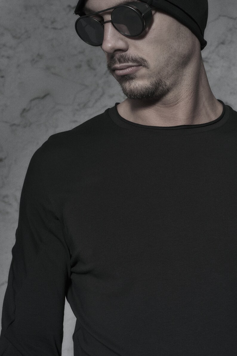 May include: A man wearing a black beanie and black sunglasses with round lenses. He is wearing a black long-sleeved shirt.