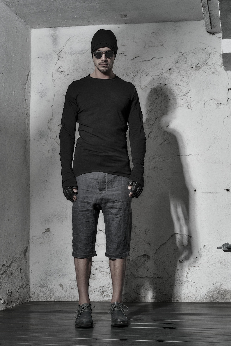 May include: A man wearing a black beanie, black sunglasses, a black long-sleeved shirt, gray shorts, and black shoes. He is standing in front of a white wall.