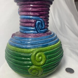 May include: A ceramic vase with a green, blue, and purple striped design. The vase has a round body and a narrow neck. The stripes are raised and have a textured finish. There are two green swirls on the lower portion of the vase.