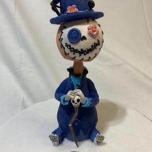 Puede incluir: Una figurita de cerámica hecha a mano de un personaje caprichoso con un sombrero de copa azul, una cara cosida con un ojo de botón y un abrigo azul que sostiene un bastón con un cráneo en la parte superior.