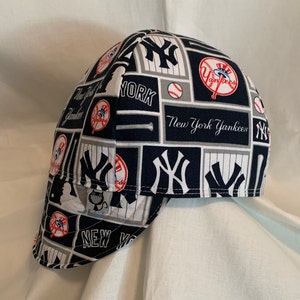 May include: A navy blue, white, and grey fabric welding cap featuring a repeating New York Yankees pattern. The design includes the team logo, baseballs, bats, and the text New York Yankees. A small grey tag with a black cat graphic is visible.