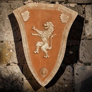 Pode incluir: Uma placa de parede em forma de escudo na cor terracota, apresentando um relevo elevado na cor creme de um leão rampante no centro. A borda é decorada com padrões esculpidos intrincados e dois motivos em forma de leque nos cantos superiores, com um pequeno emblema de cabeça de leão perto da base pontiaguda.