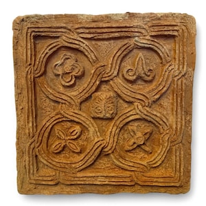 May include: A terracotta tile with a raised design of four stylized flowers in a square frame. The tile is a warm brown color and has a rough texture.