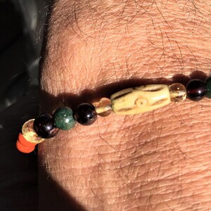 Könnte beinhalten: Ein Perlenarmband mit einer zentralen, knochenfarbenen Perle mit geätztem Design, flankiert von bernsteinfarbenen, grünen, roten und dunklen Perlen. Das Armband ist auf einer elastischen Schnur aufgefädelt. Das Armband wird an einem Handgelenk gezeigt.