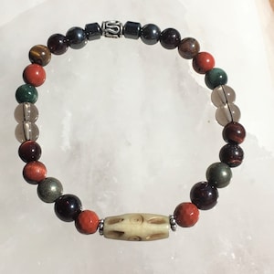 Könnte beinhalten: Ein Armband mit bunten Edelsteinen, darunter rote, grüne, braune und klare Perlen. Das Armband hat eine cremefarbene, zylindrische Mittelperle mit geätzten Mustern und einen silberfarbenen Metallakzent. Das Armband wird auf einer weißen Oberfläche präsentiert.