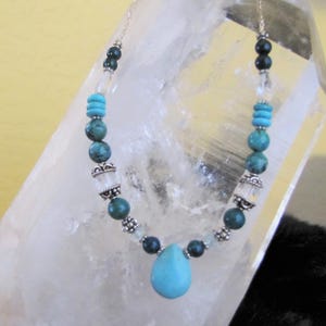 May include: A turquoise and silver necklace with a teardrop pendant. The necklace features turquoise, dark green, and clear beads, with silver accents. The necklace is displayed against a clear crystal.