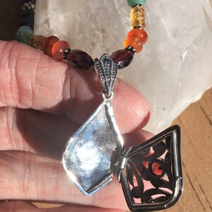 May include: A silver locket necklace with a filigree design and a red gemstone inside. The necklace is made of silver beads and features a variety of gemstones in different colors.