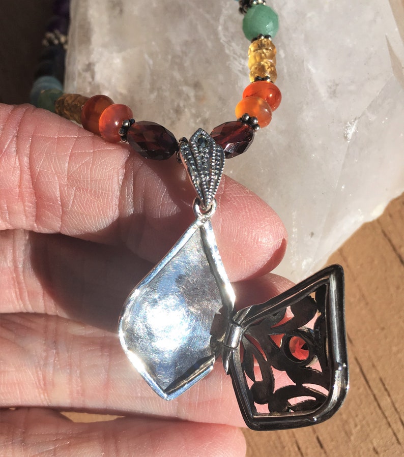 May include: A silver locket necklace with a filigree design and a red gemstone inside. The necklace is made of silver beads and features a variety of gemstones in different colors.
