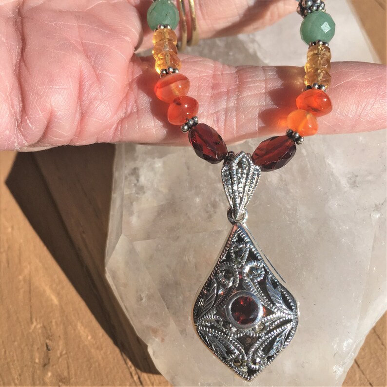 May include: A silver necklace with a teardrop-shaped pendant featuring a red garnet surrounded by silver filigree. The necklace is made with green, orange, and red beads.