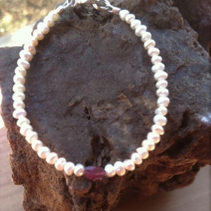 May include: A white pearl bracelet with a single red gemstone accent. The bracelet is made of small, round pearls and has a silver clasp.