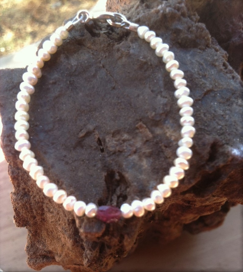 May include: A white pearl bracelet with a single red gemstone accent. The bracelet is made of small, round pearls and has a silver clasp.