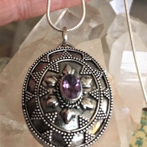 May include: A silver locket necklace with an intricate design and a purple amethyst gemstone in the center.