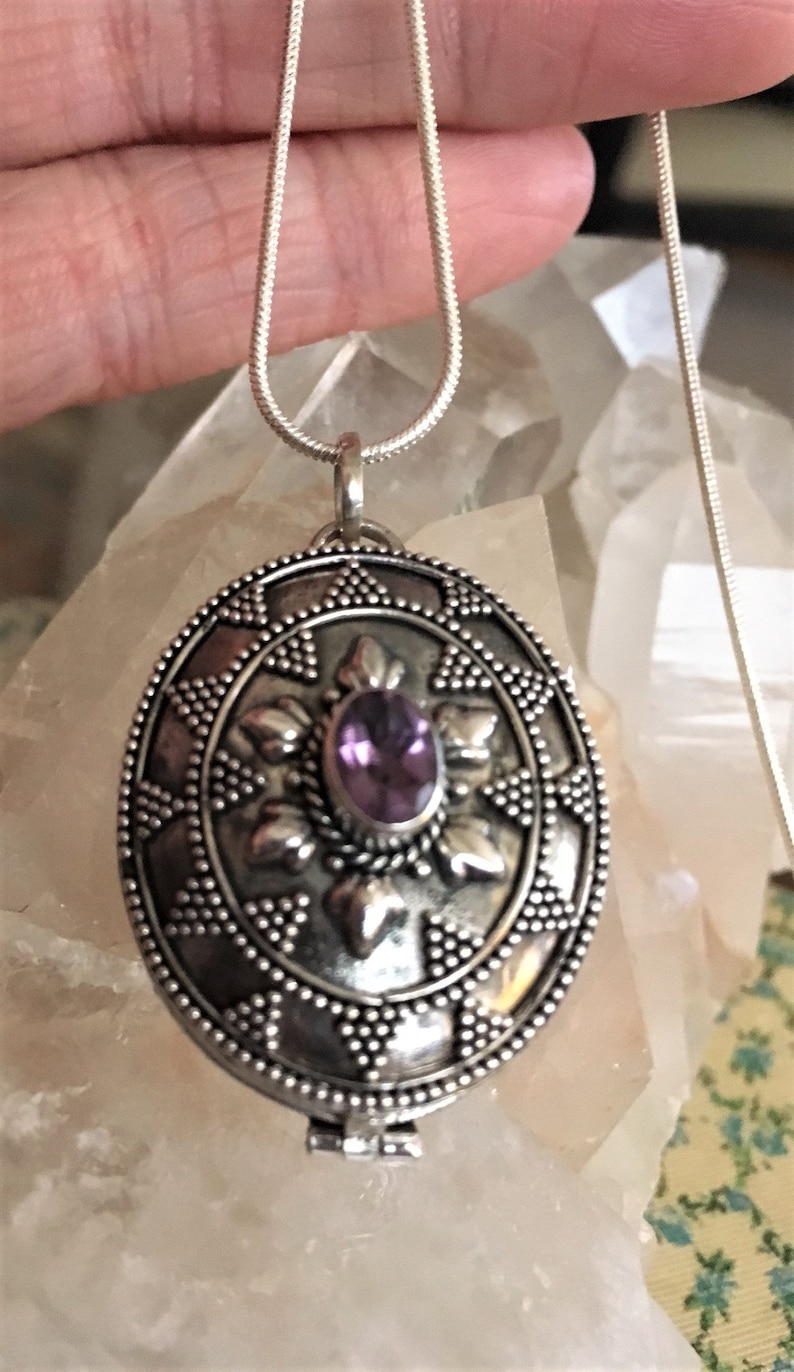 May include: A silver locket necklace with an intricate design and a purple amethyst gemstone in the center.