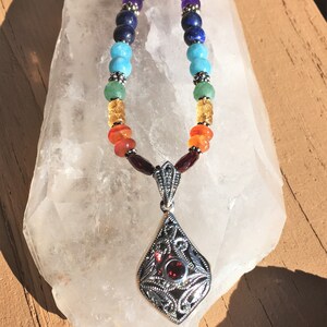 May include: A silver necklace with a pendant featuring a red gemstone surrounded by silver filigree. The necklace is made with colorful beads in a rainbow pattern.