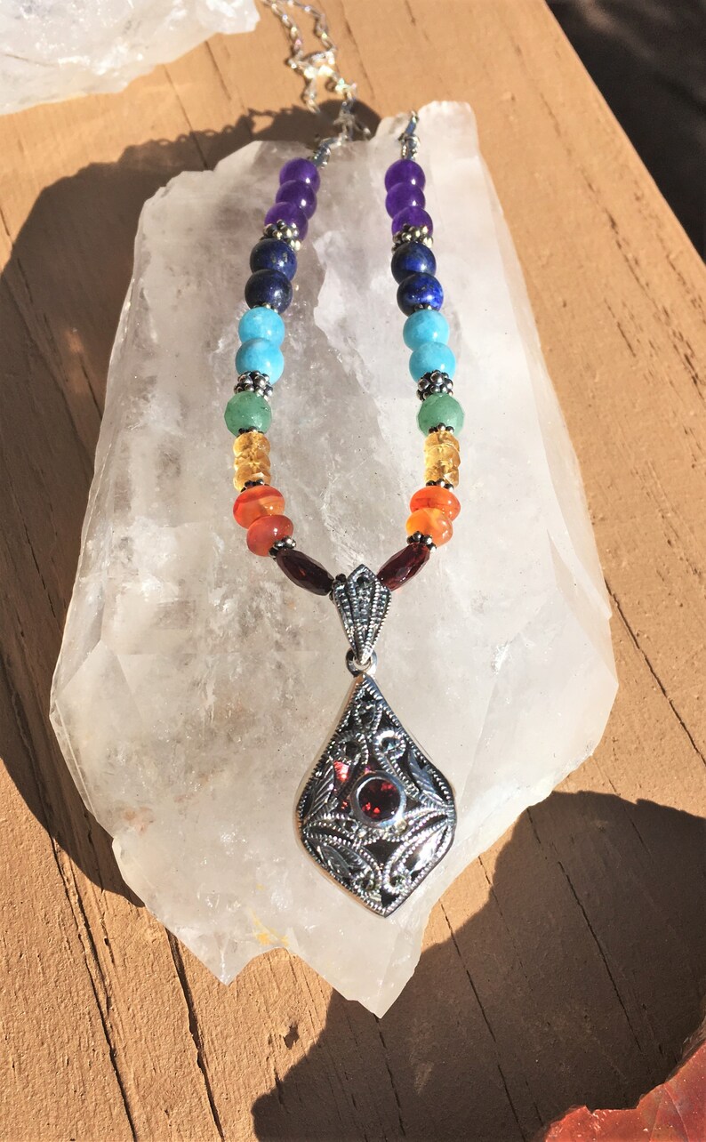 May include: A silver necklace with a pendant featuring a red gemstone surrounded by silver filigree. The necklace is made with colorful beads in a rainbow pattern.