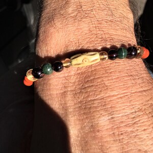 Könnte beinhalten: Ein Armband mit roten, orangefarbenen, gelben, grünen, schwarzen und braunen Perlen. Das Armband hat einen Knochenanhänger mit einem geschnitzten Design.
