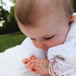 May include: A baby wearing a white lace dress with a pearl and cross bracelet. The baby is looking down with their hands clasped together.