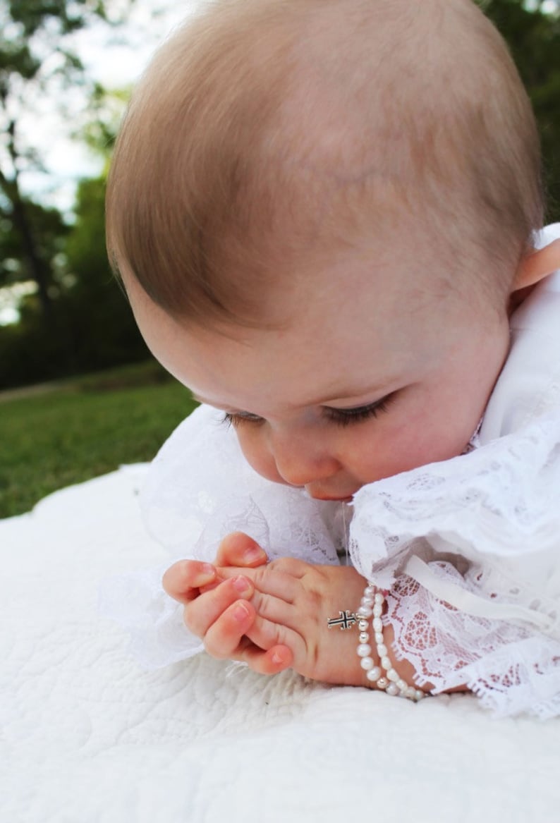 May include: A baby wearing a white lace dress with a pearl and cross bracelet. The baby is looking down with their hands clasped together.