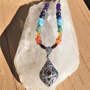 May include: A silver necklace with a pendant featuring a red gemstone surrounded by a filigree design. The necklace is made with seven different colored beads, representing the seven chakras.