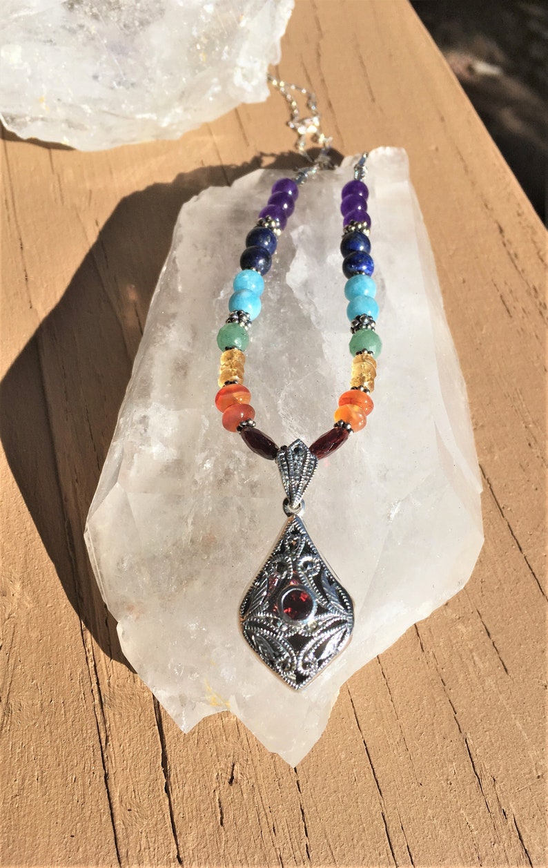 May include: A silver necklace with a pendant featuring a red gemstone surrounded by a filigree design. The necklace is made with seven different colored beads, representing the seven chakras.