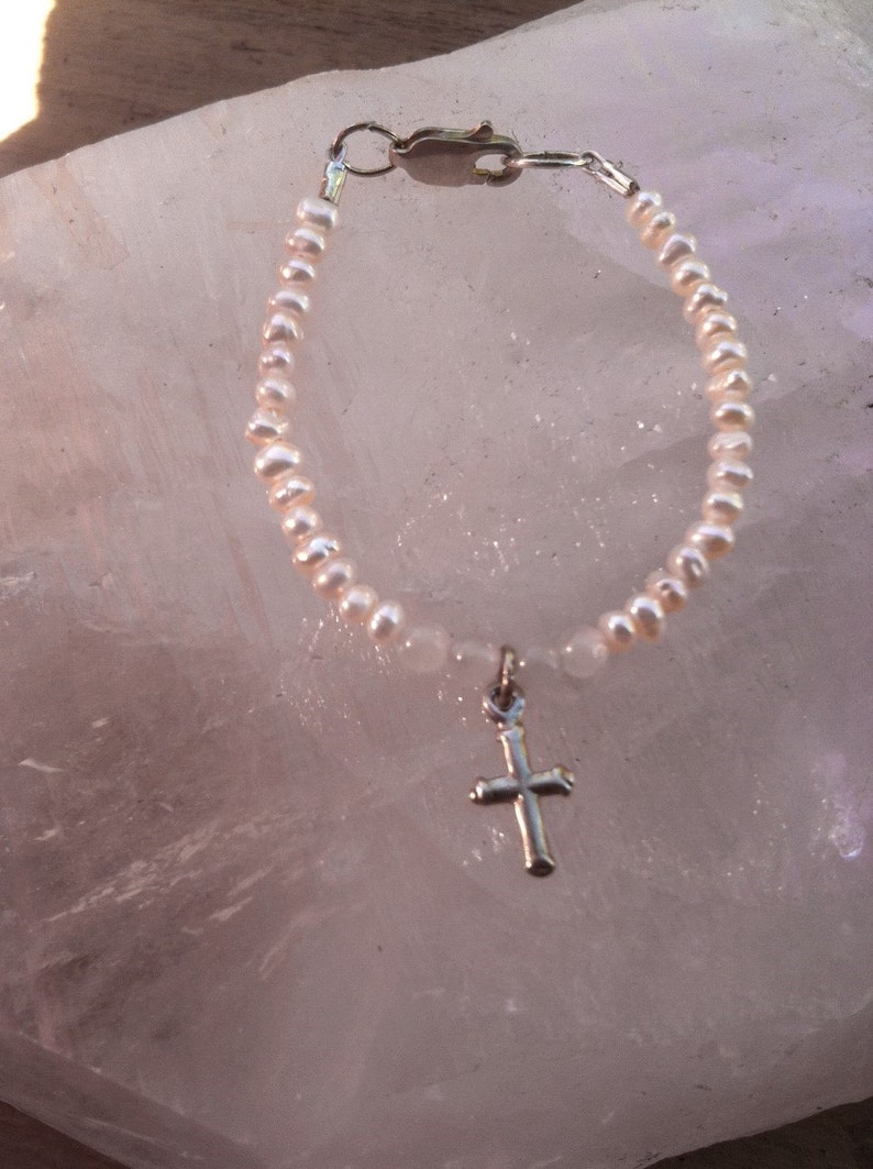 May include: A delicate pearl bracelet with a silver cross charm. The bracelet is made of small, white pearls and has a simple, elegant design.