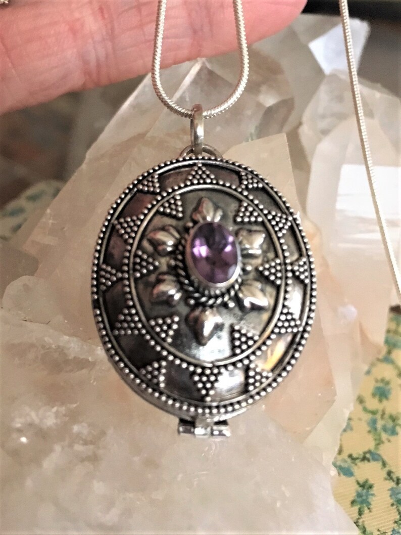 May include: A silver locket necklace with an intricate design and a purple gemstone in the center.